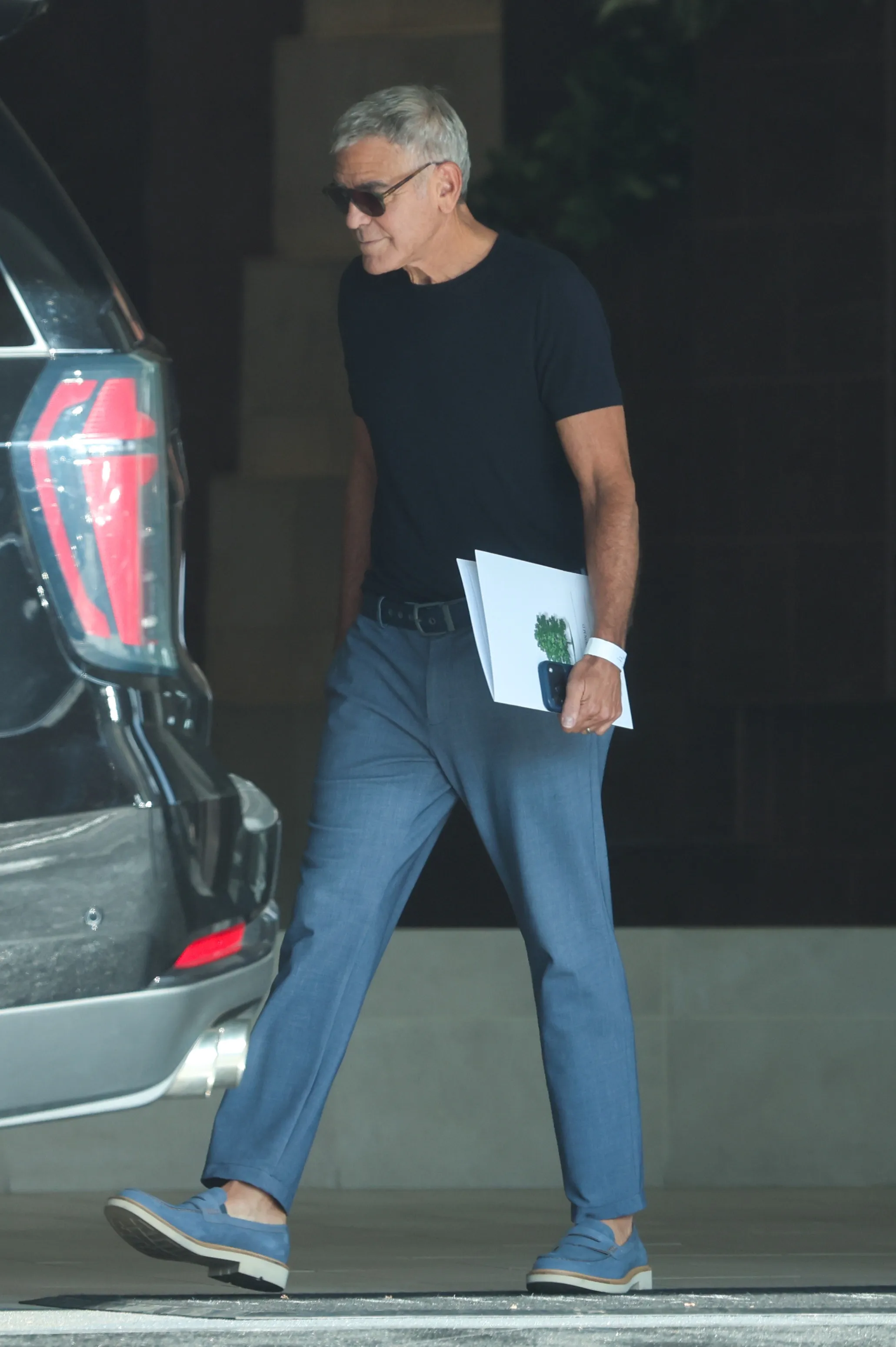 George Clooney wearing sunglasses, a black t-shirt, and blue trousers, with a bandage on his right hand, carries paperwork toward a vehicle.