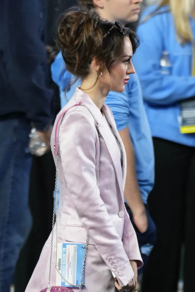 Jordon Hudson wearing a pink blazer and carrying a chain-strap bag on the field.