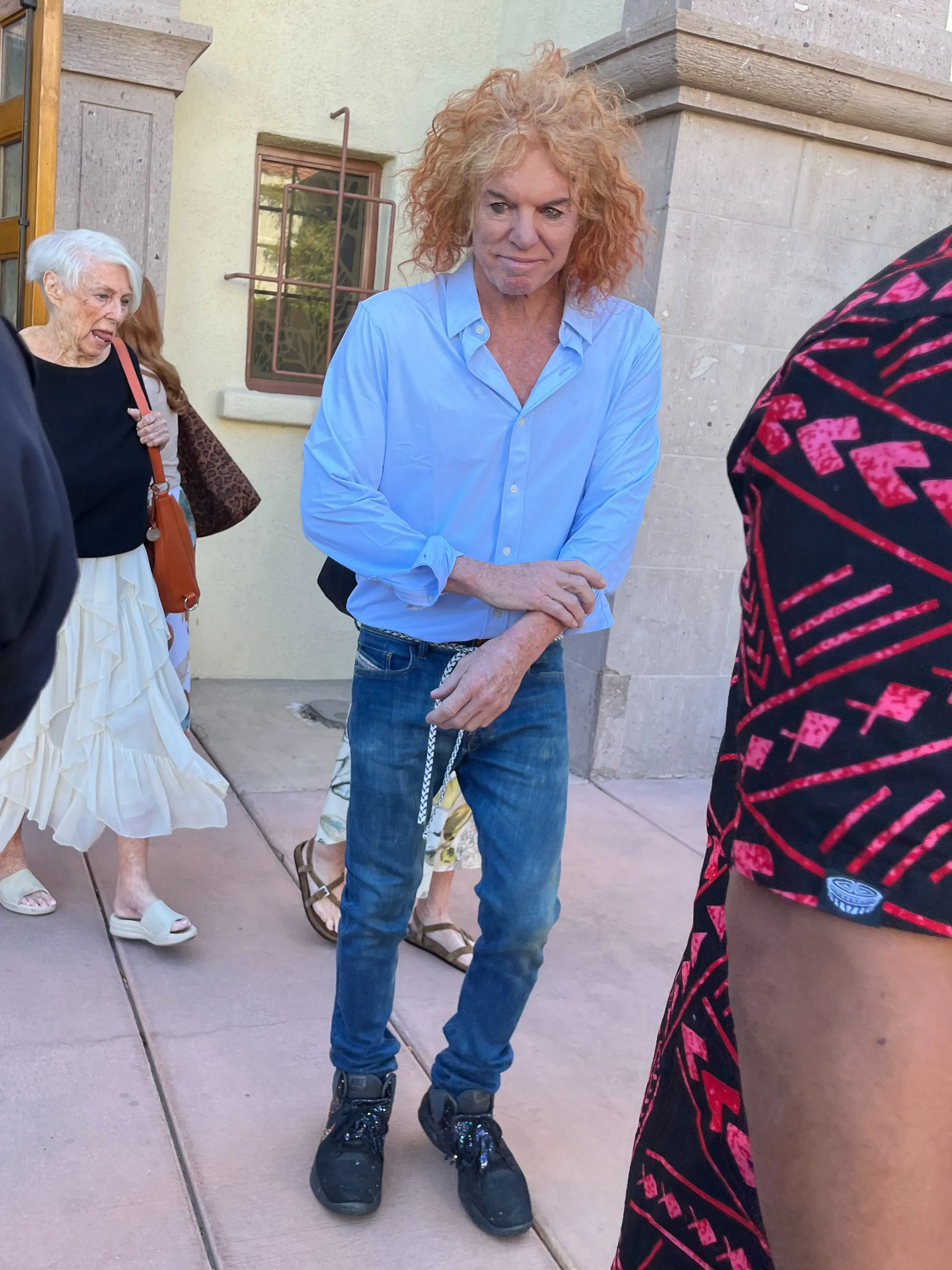 Comedian Carrot Top wearing a light blue button-down shirt and blue jeans stands outside a building.