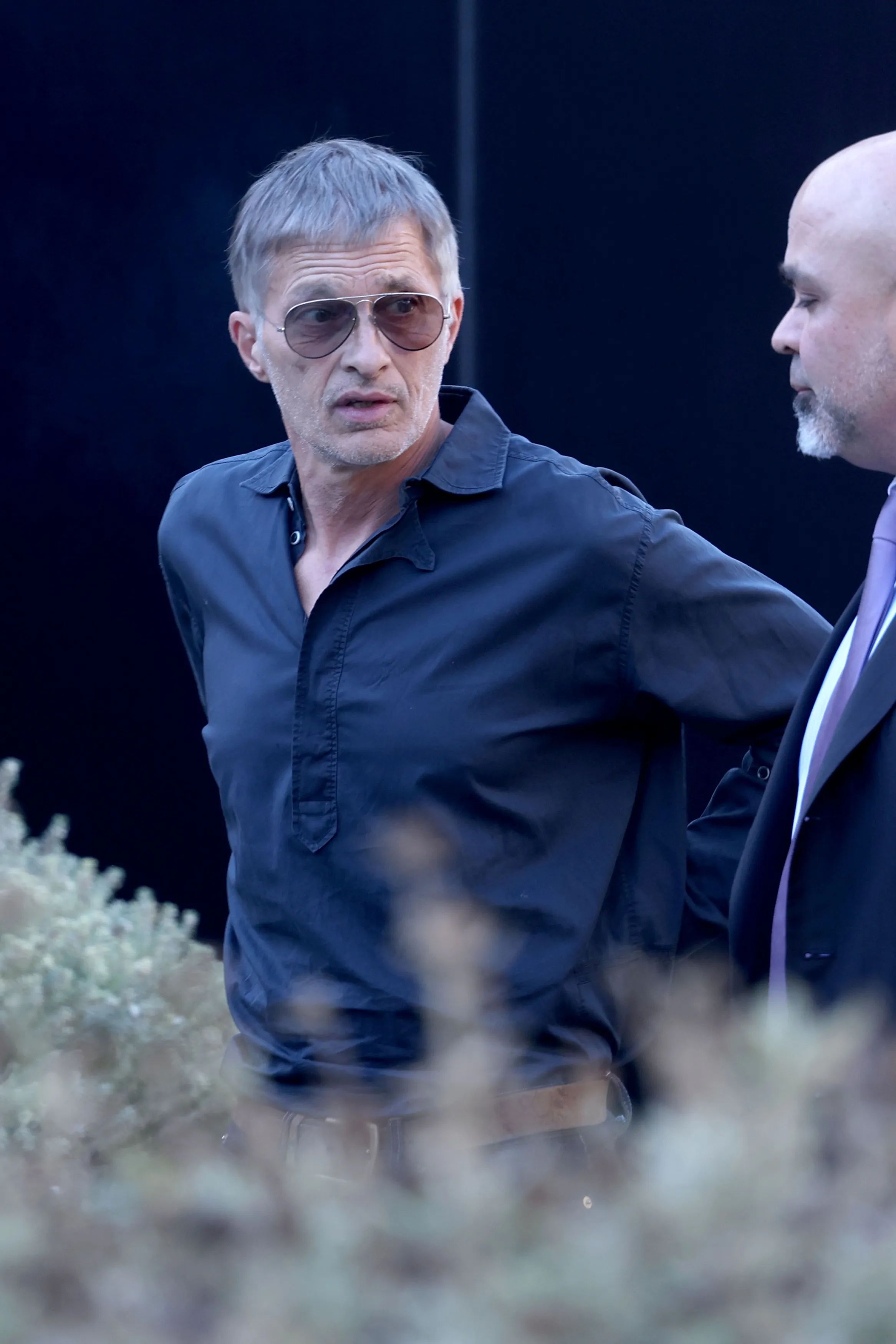 Olivier Martinez wearing sunglasses and a dark shirt, standing near a lawyer outside a courthouse.