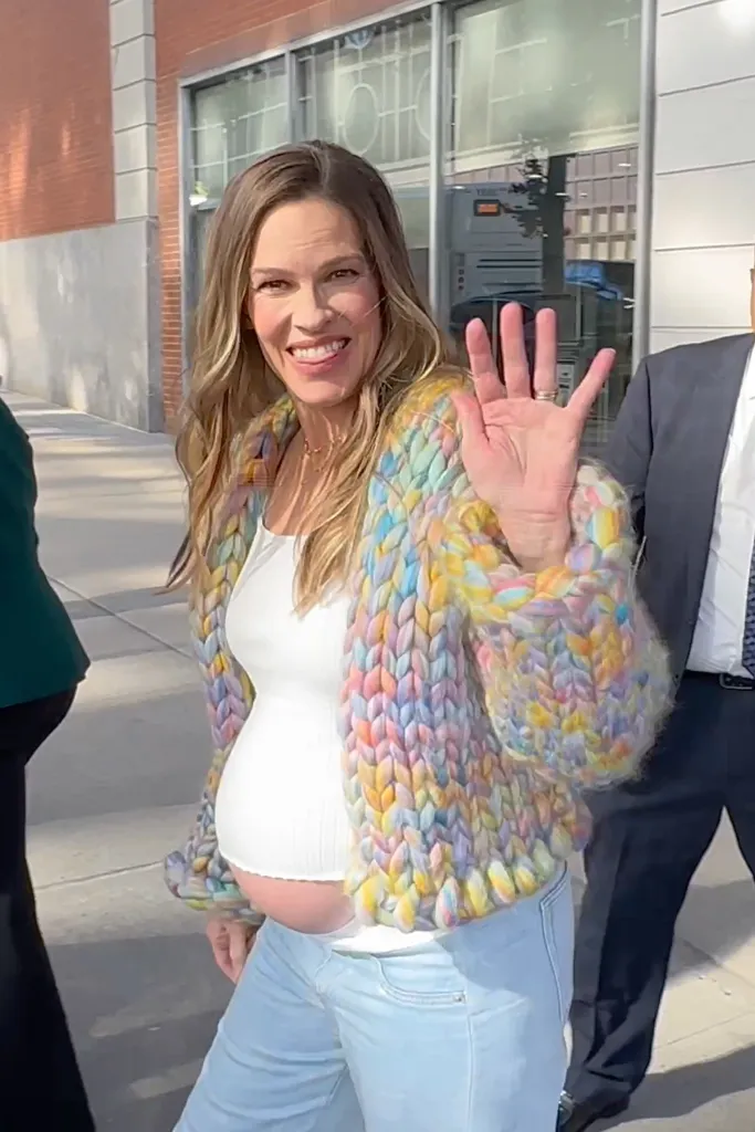 Hilary Swank smiles and waves to fans outside CBS Studios in New York City, wearing a white top and a pastel cardigan.