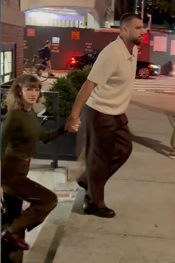 Taylor Swift and Travis Kelce hold hands while walking down a street at night.