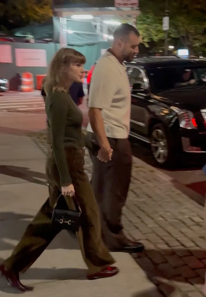 Taylor Swift and Travis Kelce hold hands while walking down a city sidewalk at night.