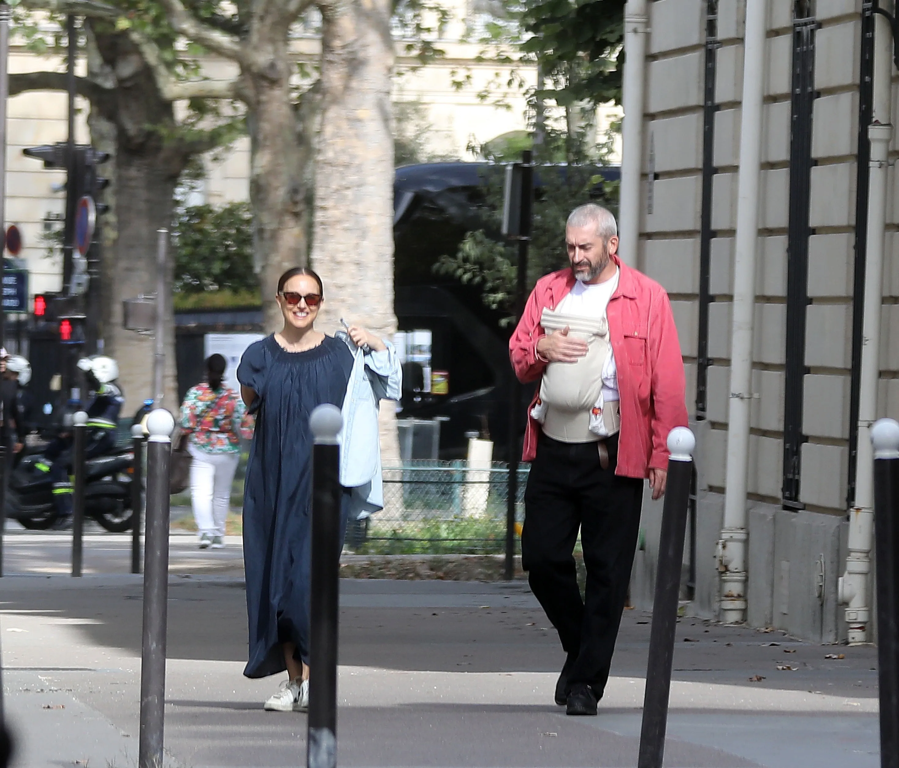 Natalie Portman and her partner Tanguy walk down a Paris street, with Tanguy carrying their newborn baby in a carrier.
