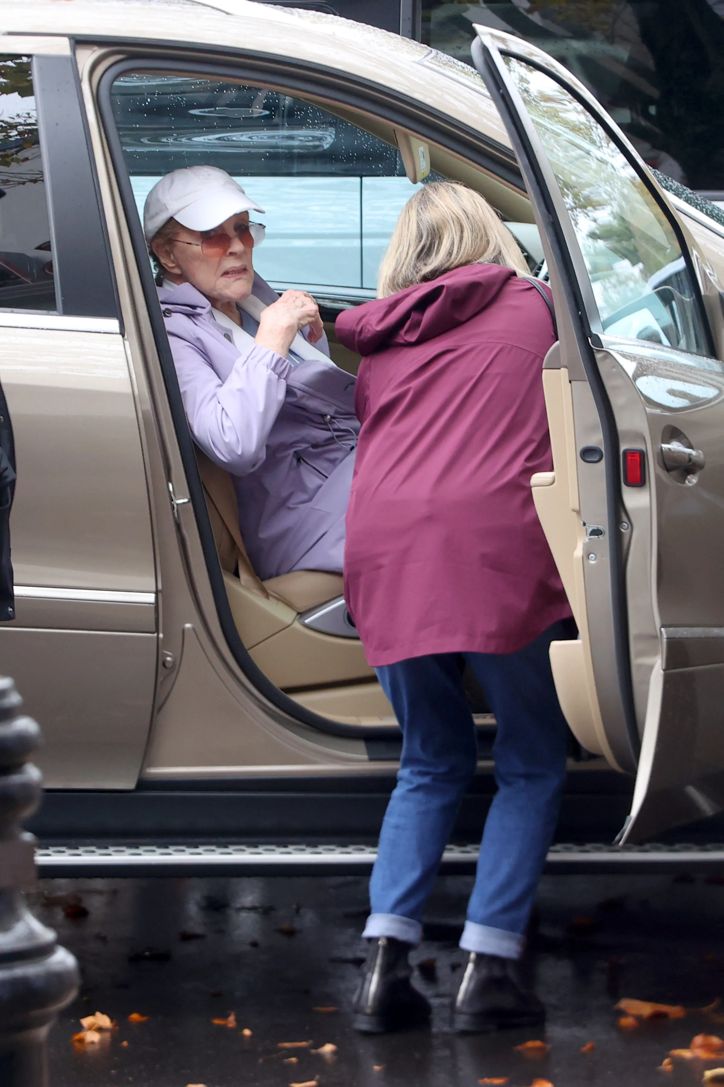 Julie Andrews sits in the passenger seat of a car, wearing a white baseball cap, sunglasses, and a purple jacket.