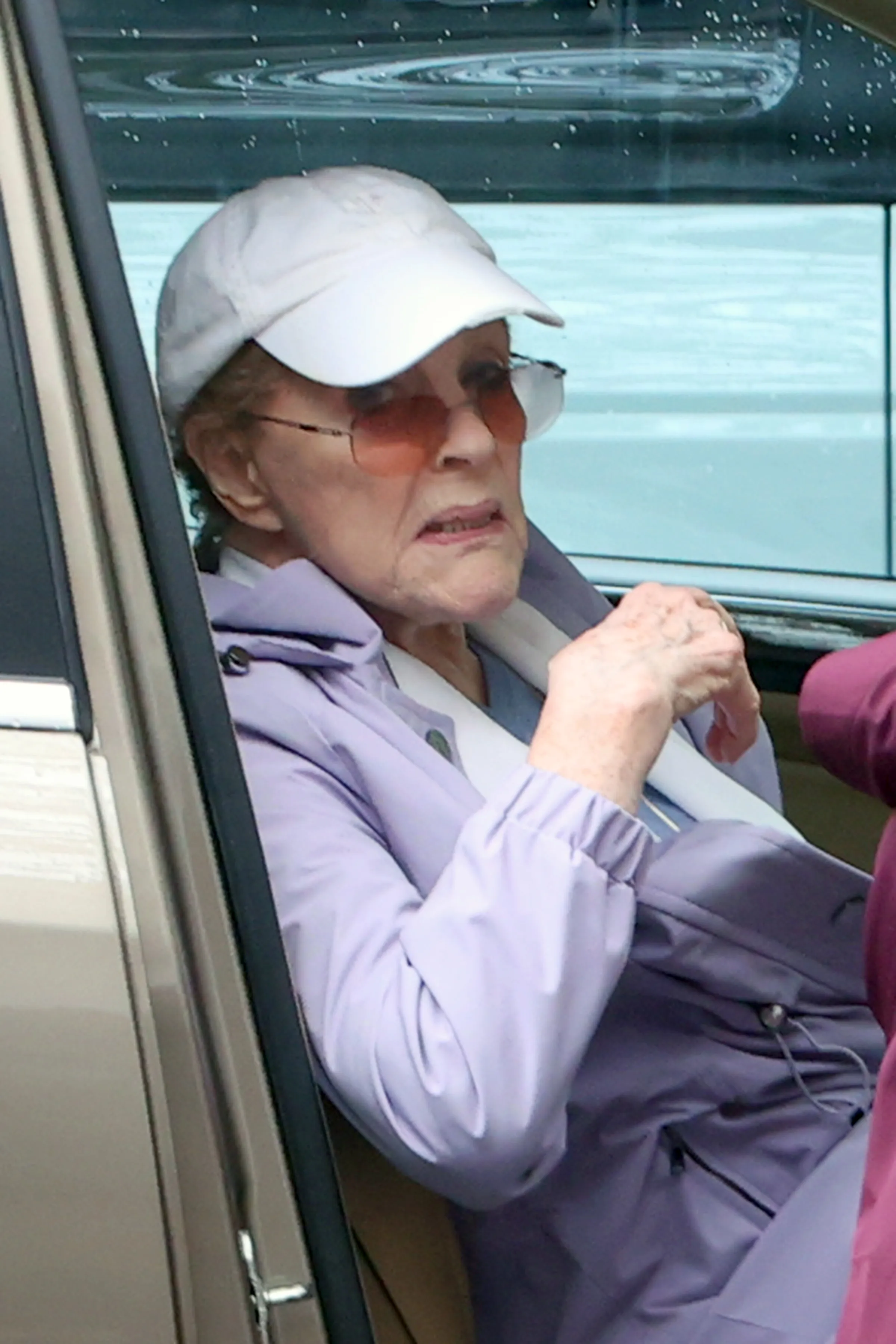 Julie Andrews wearing a white baseball cap and sunglasses in the passenger seat of a car.