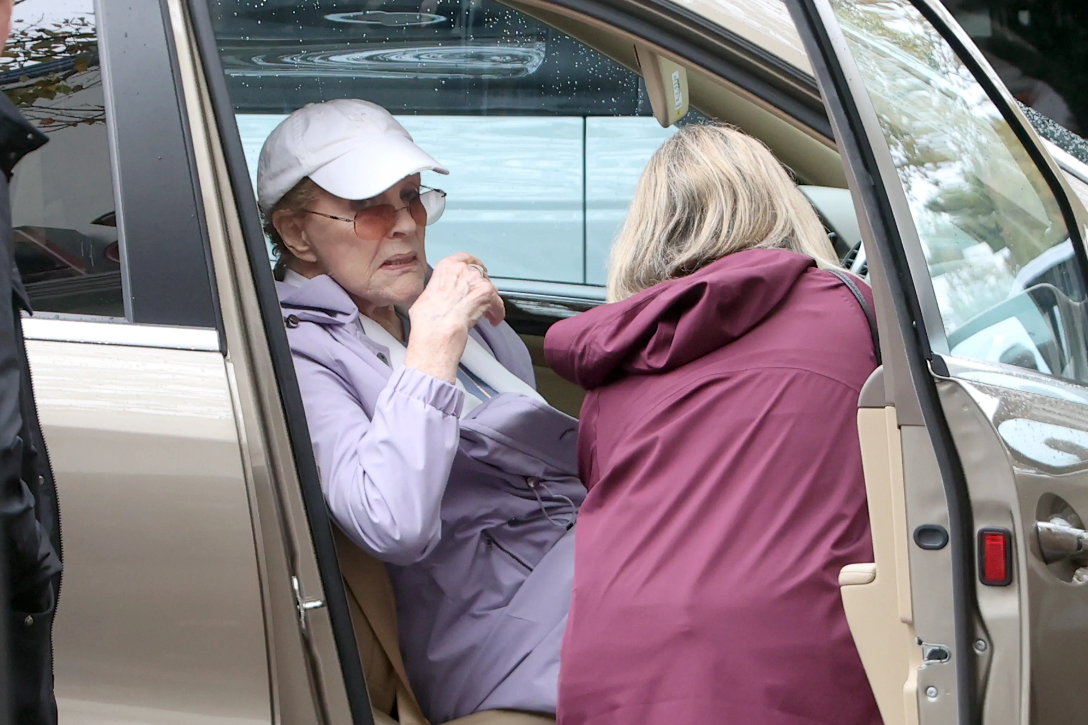 Julie Andrews seated in the passenger seat of a car, wearing a white baseball cap and sunglasses.