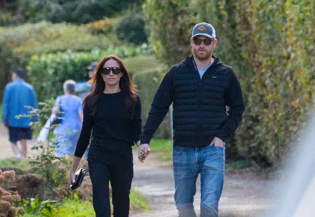 Prince Harry in a black sweater and blue jeans, and Meghan Markle, wearing a black sweater and black pants, holding hands while walking outdoors.