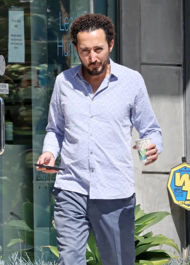 Dr. Reza Nabavi walks outside, wearing a light blue button-down shirt and grey trousers, holding a smartphone and a takeaway coffee cup.