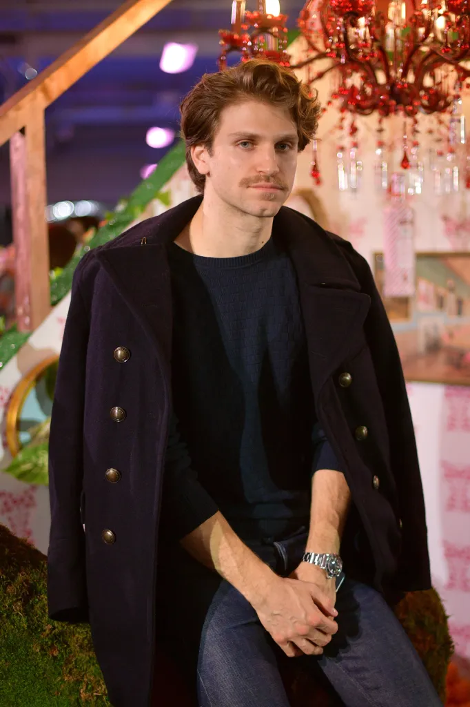 Keegan Allen wearing a dark coat draped over his shoulders and a blue sweater sits posing for the camera.