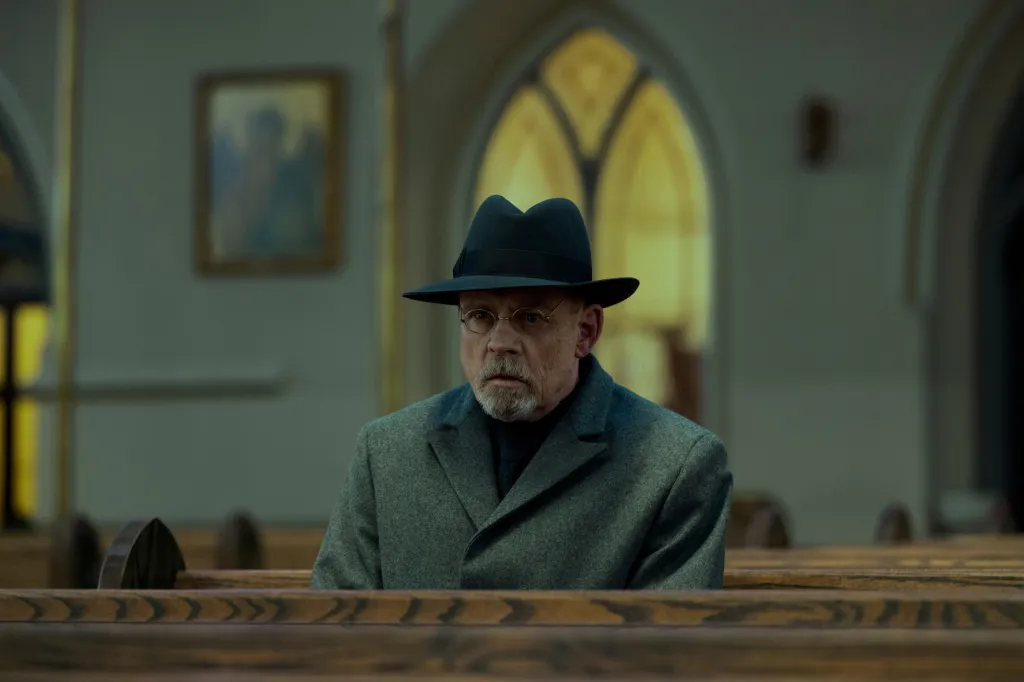 Mark Hamill as Arthur Pym in The Fall of the House of Usher, sitting in a church pew wearing a fedora and grey coat.