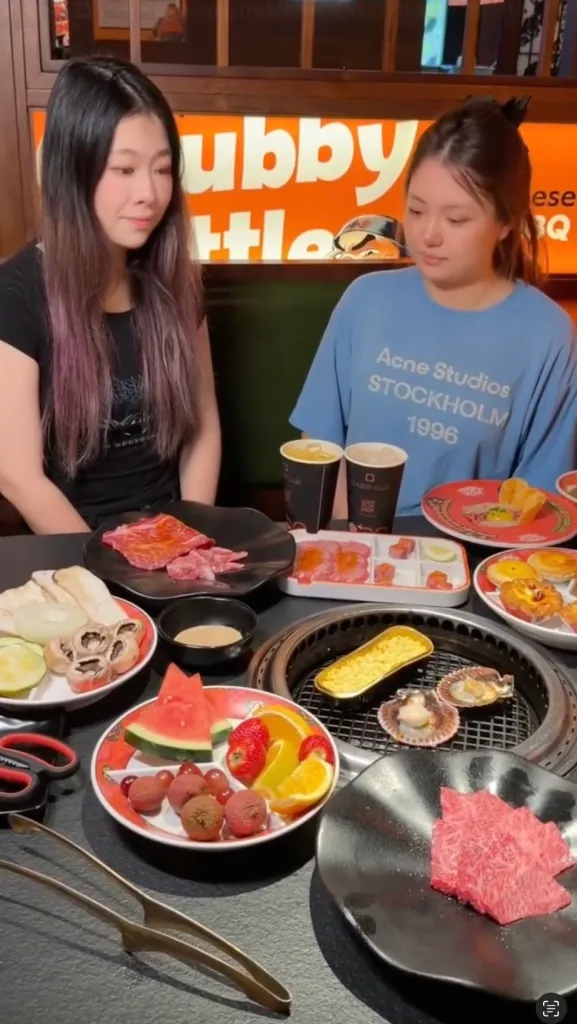 Two young women sitting at a table filled with various dishes, including raw meat, vegetables, seafood, fruit, and drinks, at a restaurant.