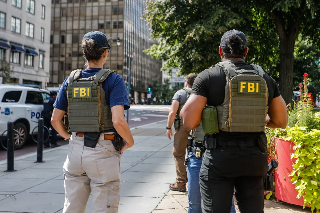An FBI agent and a Metropolitan Police Department officer stand on a city sidewalk wearing tactical vests with