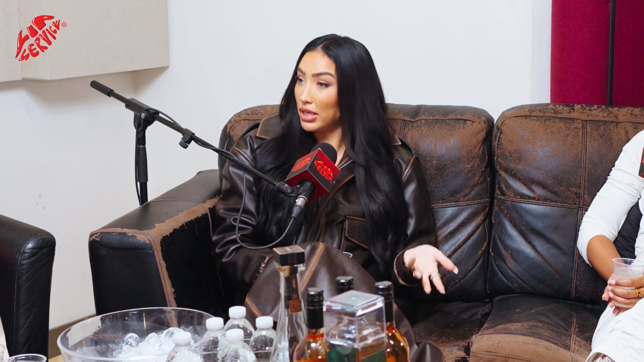 Bre Tiesi seated on a brown leather couch in a podcast studio, speaking into a microphone.