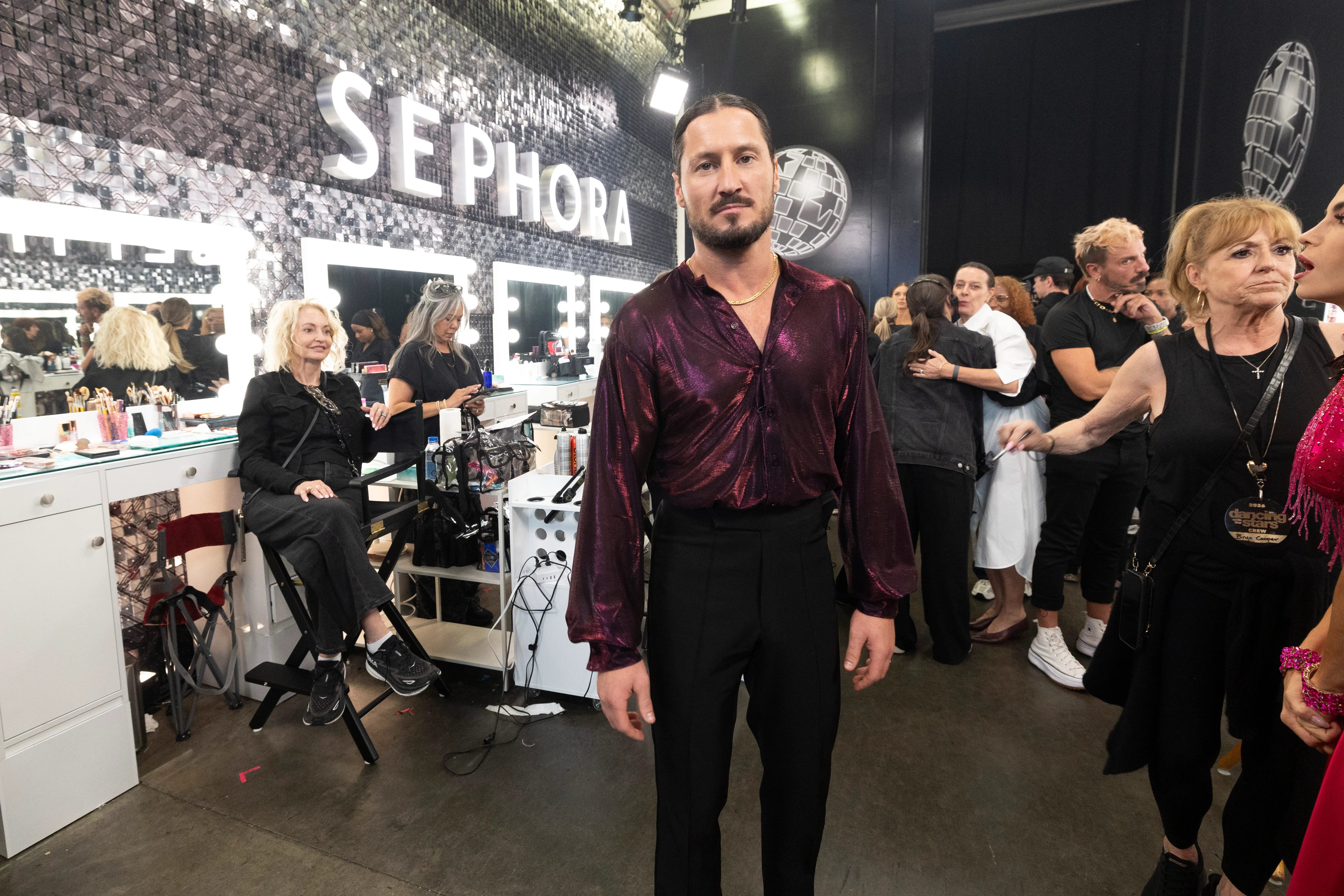 Valentin Chmerkovskiy standing backstage in a dressing room, wearing a shiny purple shirt and black pants.