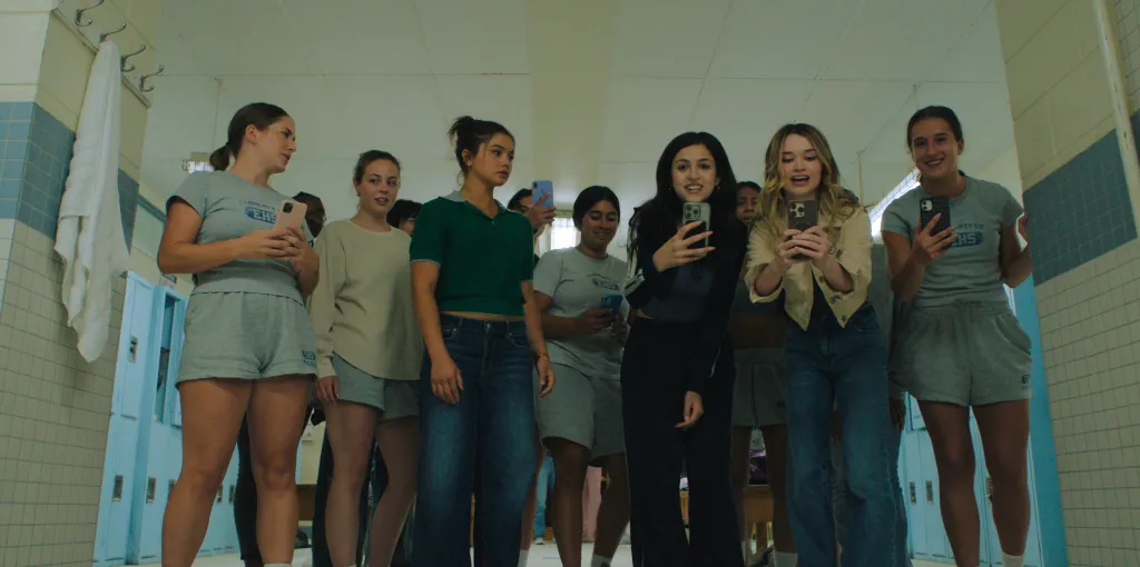 A group of young women stand in a locker room, looking down while holding up their smartphones.