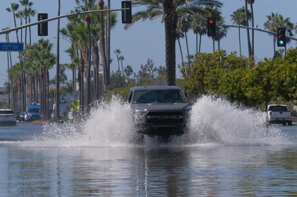 A gray pickup truck splashes through floodwaters covering a street lined with palm trees on the Balboa Peninsula in Newport Beach, California.