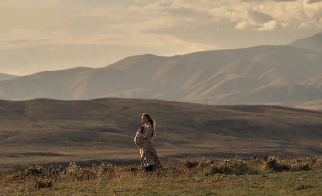 Florence Pugh stands in a striped dress against a vast mountainous landscape.