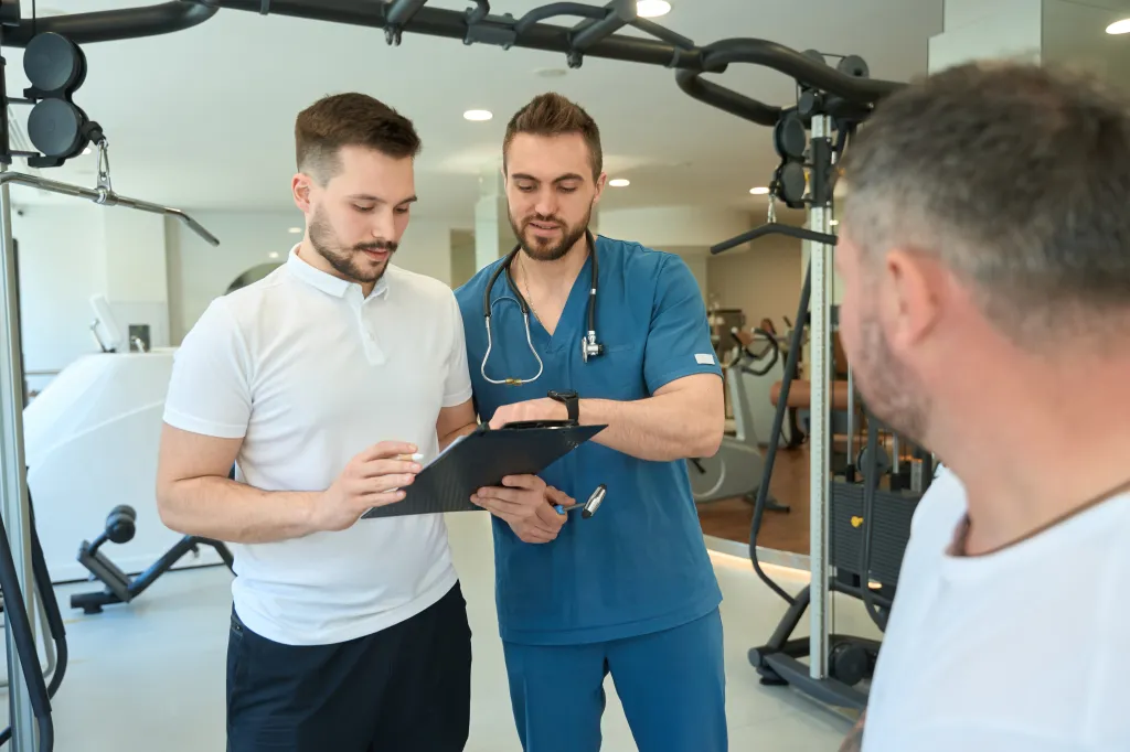A physiotherapist shows a physical rehabilitation plan to a fitness coach in the presence of a client.