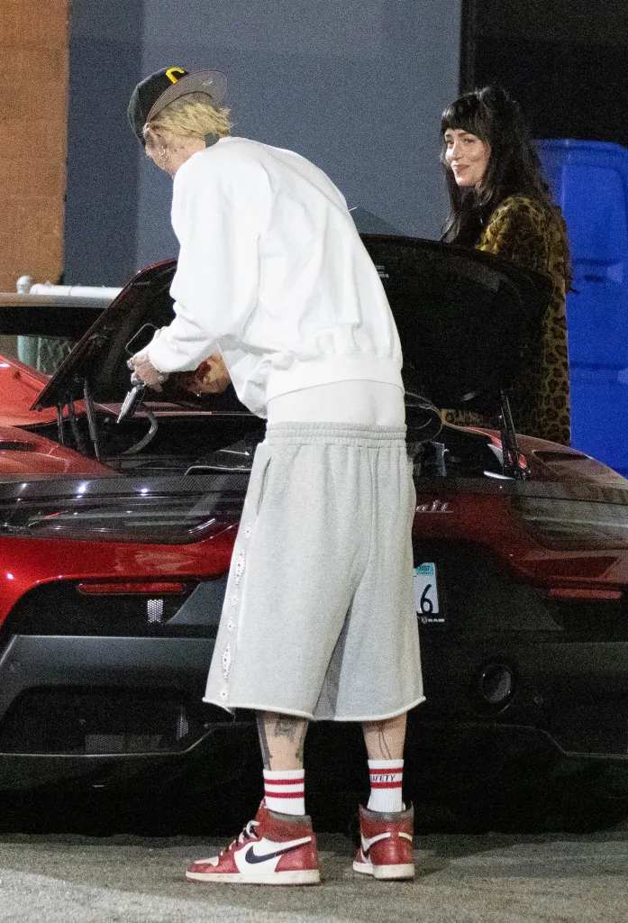 Dakota Johnson stands by a red car while Machine Gun Kelly cleans out the trunk.