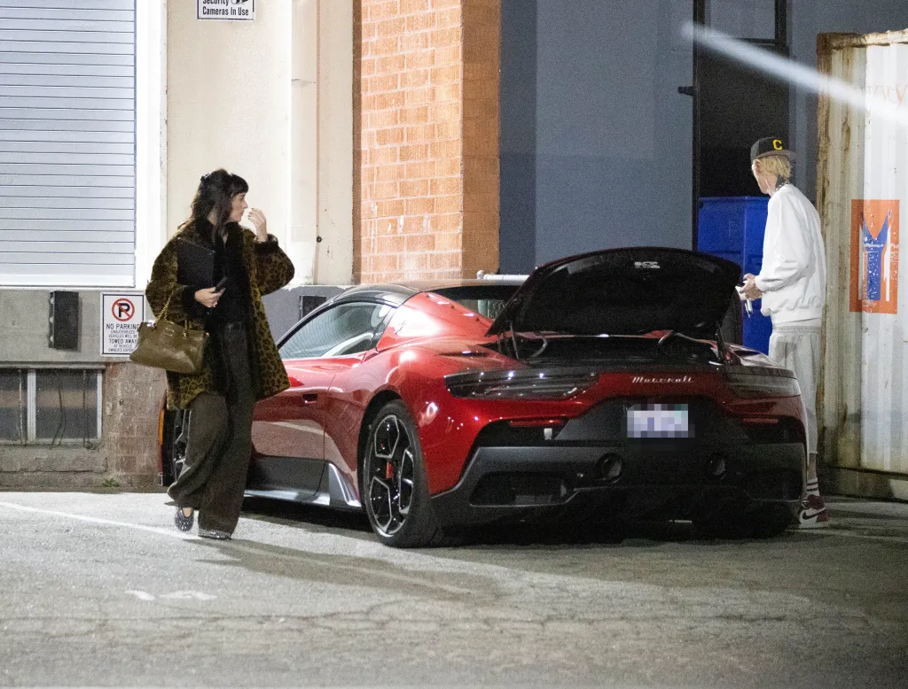 Dakota Johnson waits by a red sports car as Machine Gun Kelly stands nearby in a parking lot.
