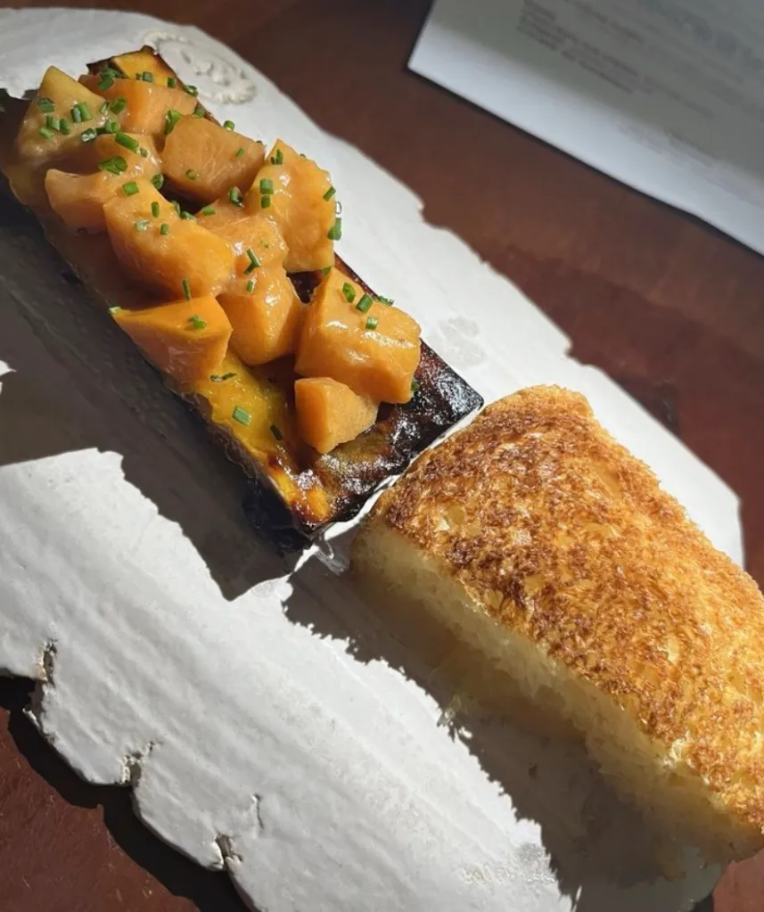 A fine-dining dish made by Michael Warring consisting of roasted bone marrow topped with diced orange fruit and chives, served with a piece of toasted bread.