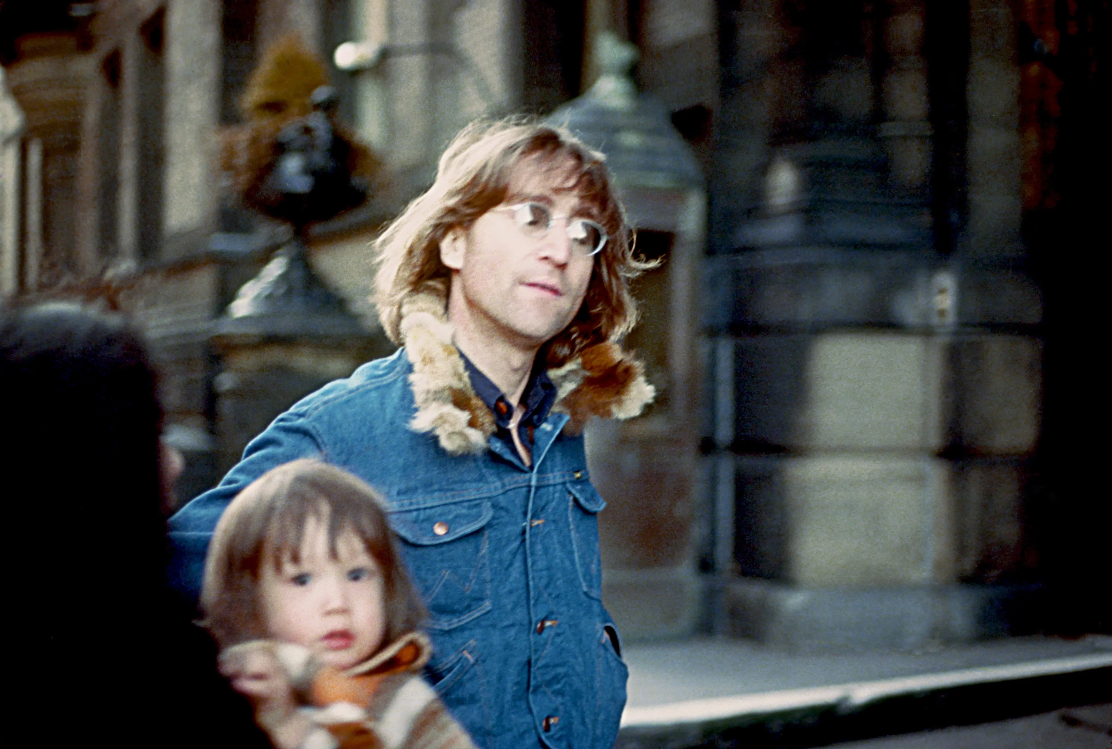 John Lennon wearing a denim jacket with a fur collar, walking with his young son Sean in New York City.
