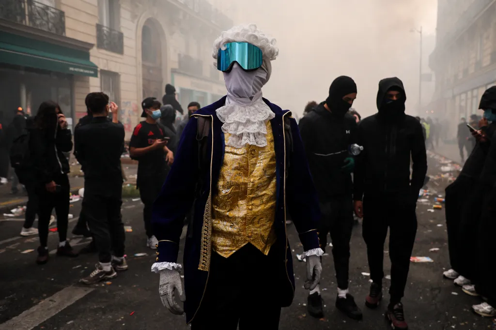 A protester in an 18th-century costume, powdered wig, and reflective ski goggles stands amid a cost-of-living demonstration in Paris.