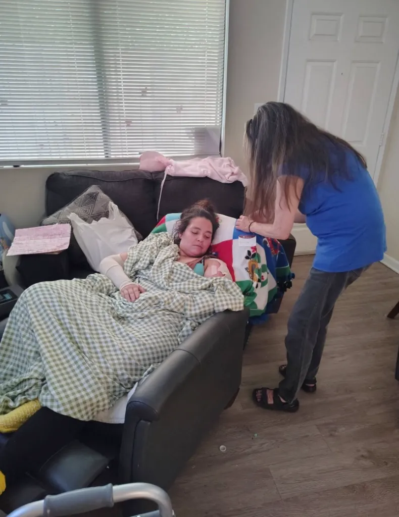 Frances Lopez resting in a reclining chair, covered in a blanket with bandages on her arm, while another person stands nearby.