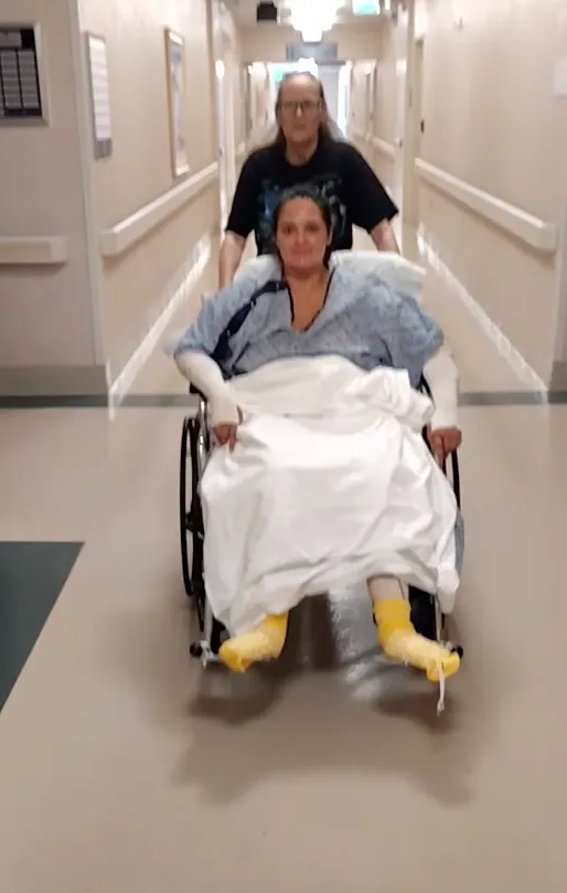 Frances Lopez in a wheelchair in a hospital hallway, with another person pushing her from behind.