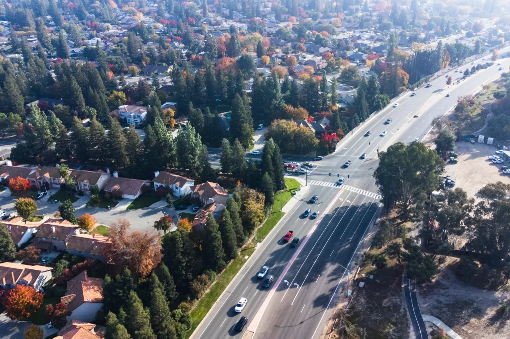 Aerial view of suburban development in Fresno, California, showing houses, trees with autumn foliage, and a multi-lane road with cars.