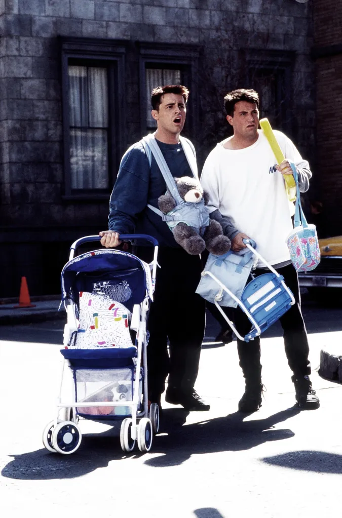 Matt LeBlanc and Matthew Perry standing outside on a street holding baby accessories, pushing a stroller and carrying baby bags from the television show Friends.