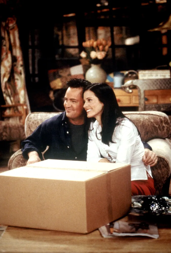 Matthew Perry and Courteney Cox sitting on a couch behind a cardboard box in the television show Friends.