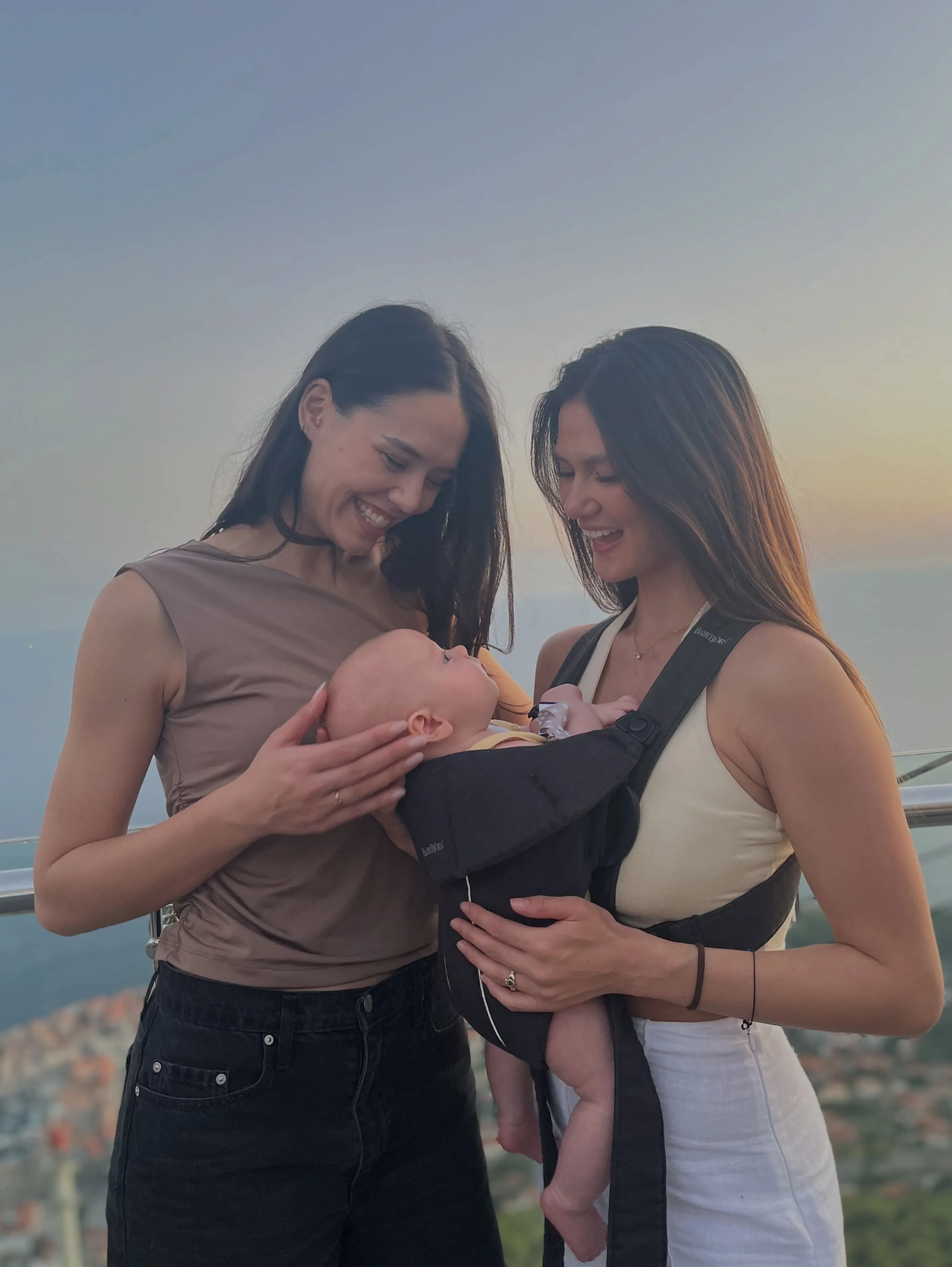 Two women smiling down at a baby being carried in a baby carrier outdoors at sunset.
