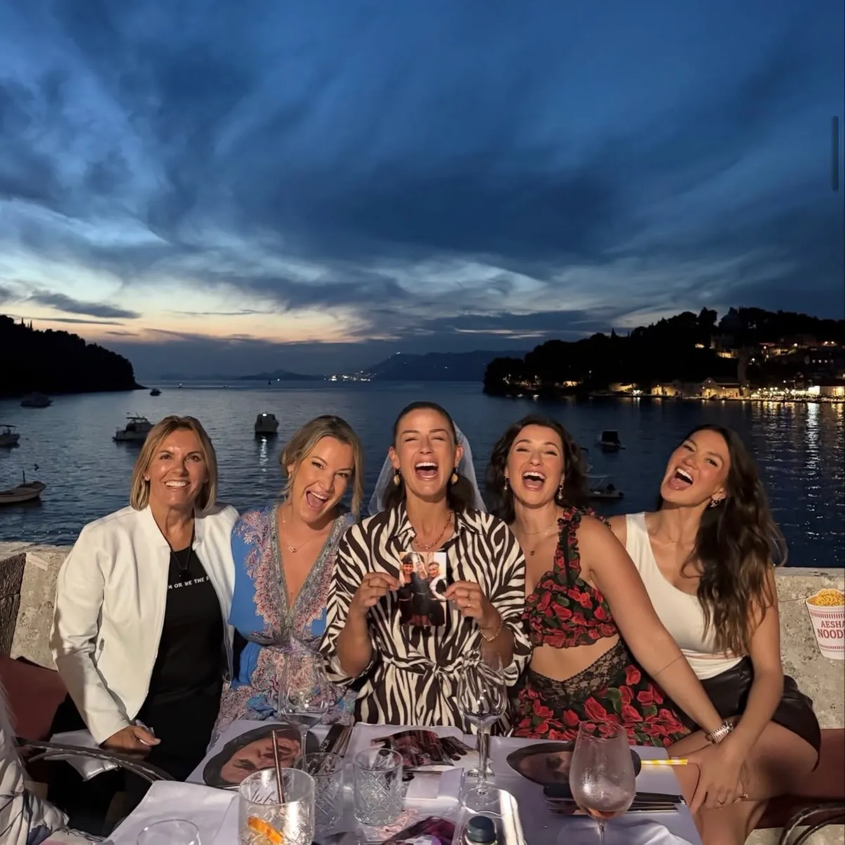 Five women laughing and celebrating at an outdoor seaside table at dusk, with the woman in the center holding up a small picture.
