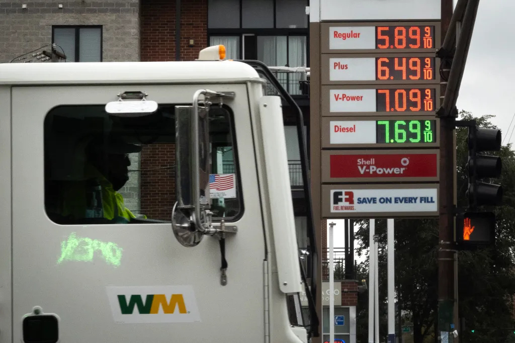 Gas station sign displaying high fuel prices with a white truck in the foreground.