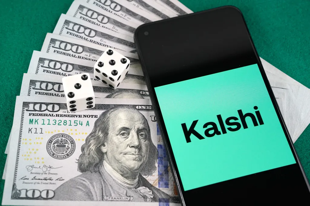 A smartphone displaying the Kalshi logo rests on a stack of hundred-dollar bills alongside two dice.