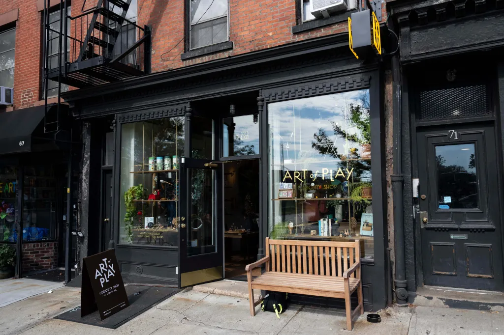 Exterior view of the Art of Play store in Brooklyn, showing its storefront, a wooden bench outside, and a sign on the sidewalk.