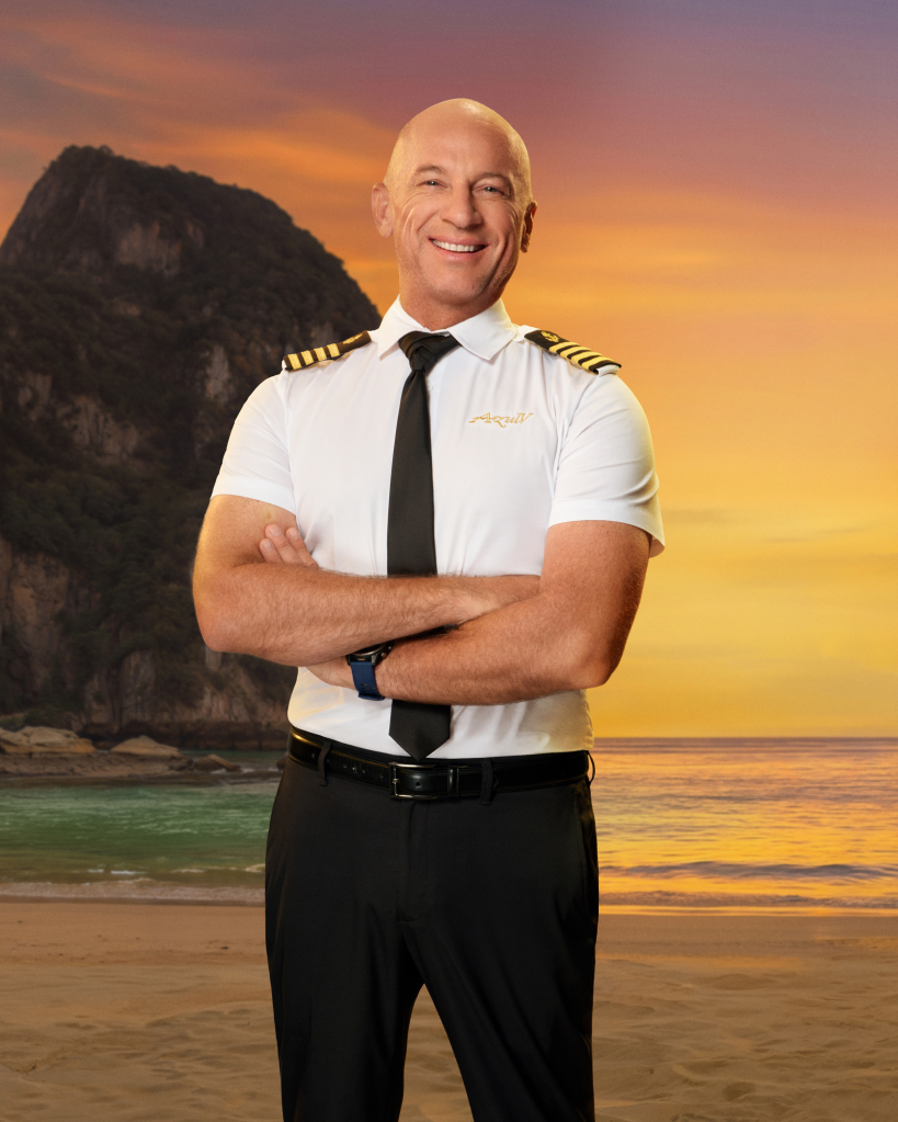 A smiling airline pilot stands with arms crossed on a beach at sunset, with a large rocky mountain in the background.