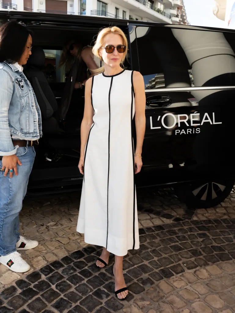 Gillian Anderson stands in front of a black L'Oréal Paris van, wearing a white and black sleeveless dress and sunglasses.