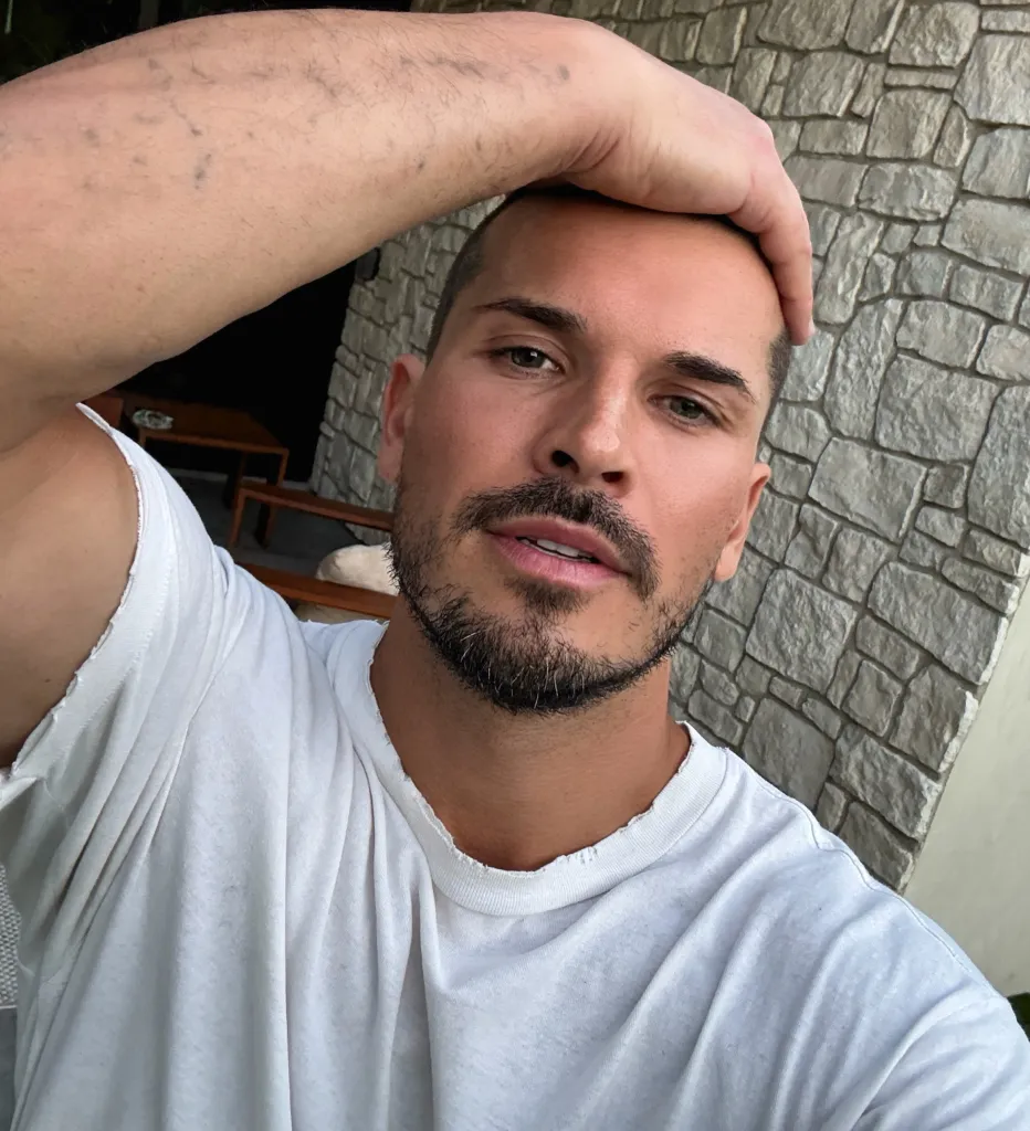 Gleb Savchenko poses for a selfie in front of a stone wall, wearing a white t-shirt and holding his hand to his head.