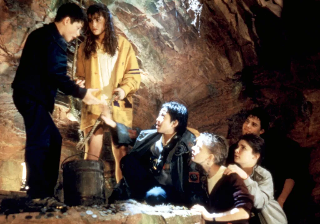 Cast members from The Goonies inside a dimly lit cave, with Sean Astin pulling a bucket on a rope and Ke Huy Quan crouching nearby.