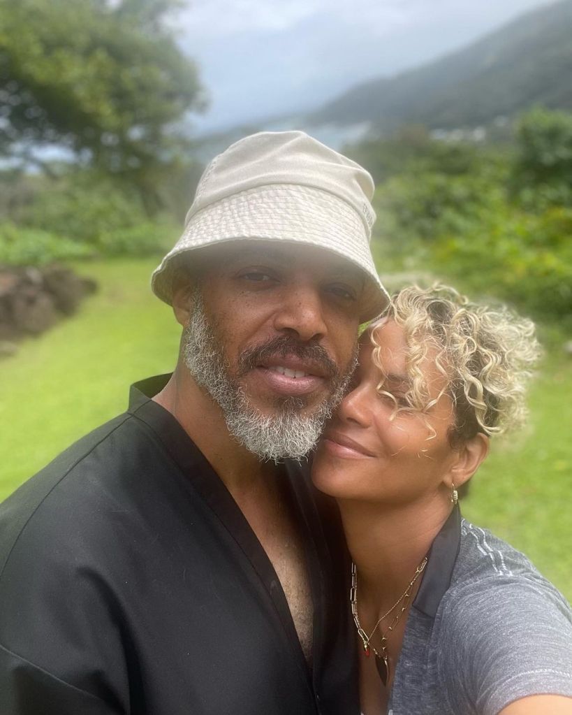 Halle Berry, in a grey T-shirt, and Van Hunt, in a black shirt and cream bucket hat, smiling outdoors with a scenic background.
