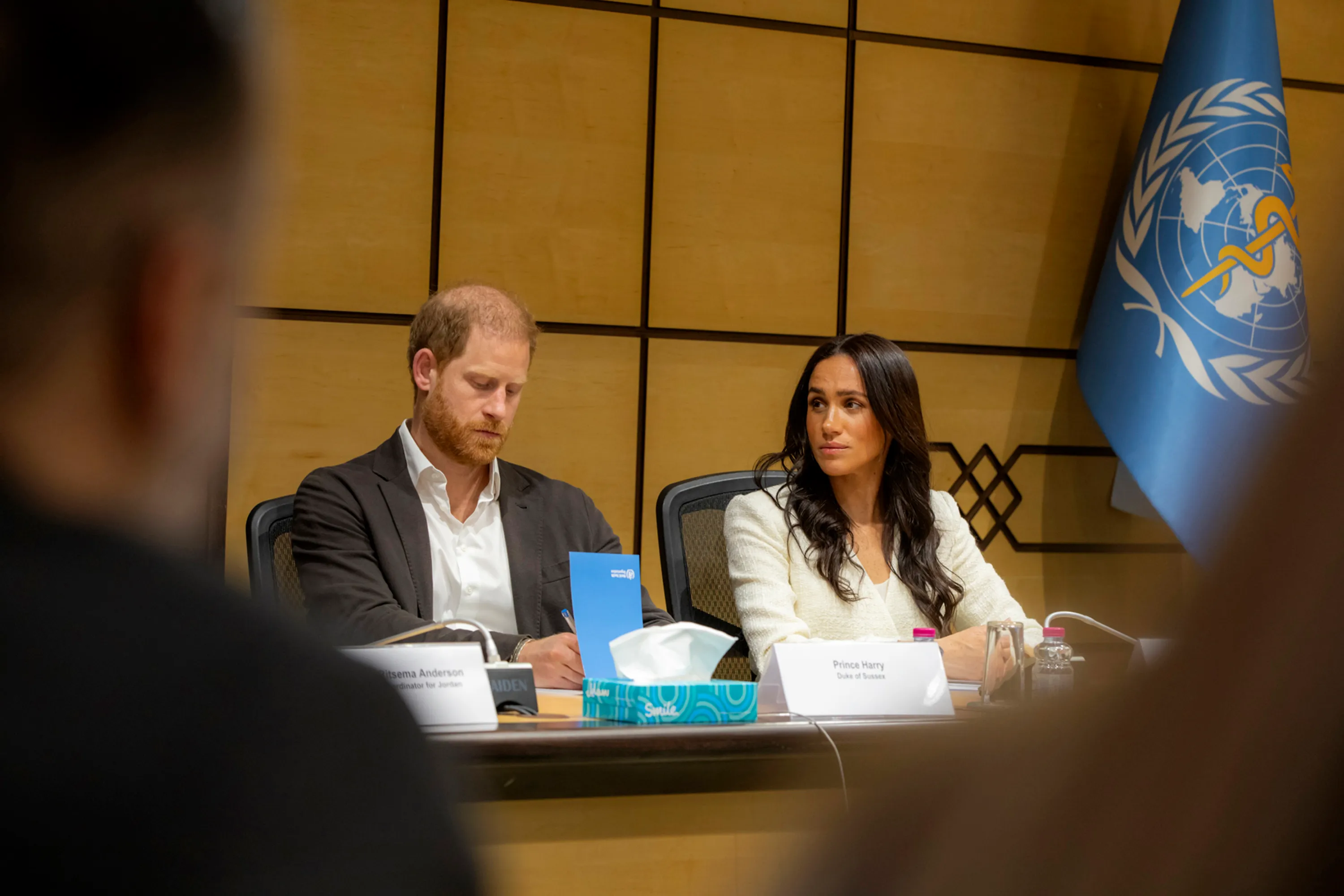 Prince Harry and Meghan Markle sit at a table during a World Health Organisation roundtable discussion in Amman, Jordan, with a WHO flag visible in the background.