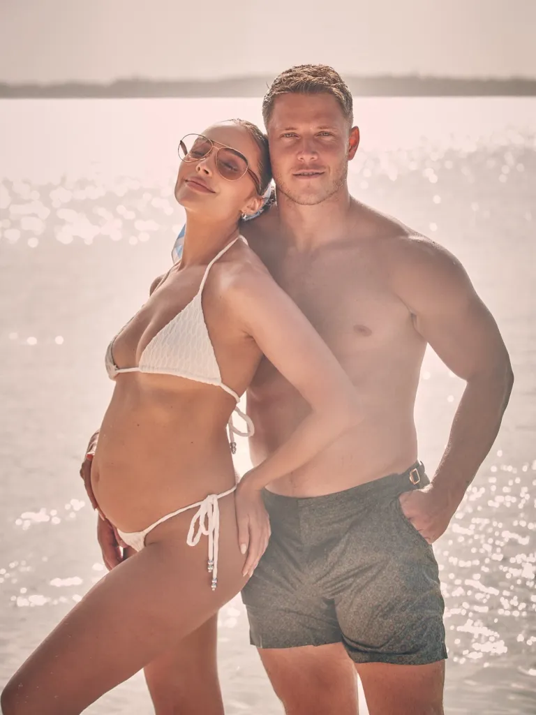 Olivia Culpo and Christian McCaffrey posing together on a beach in swimwear, with Culpo cradling her baby bump.