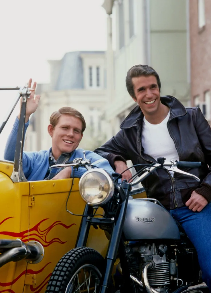 Ron Howard waving from a yellow car and Henry Winkler sitting on a motorcycle.