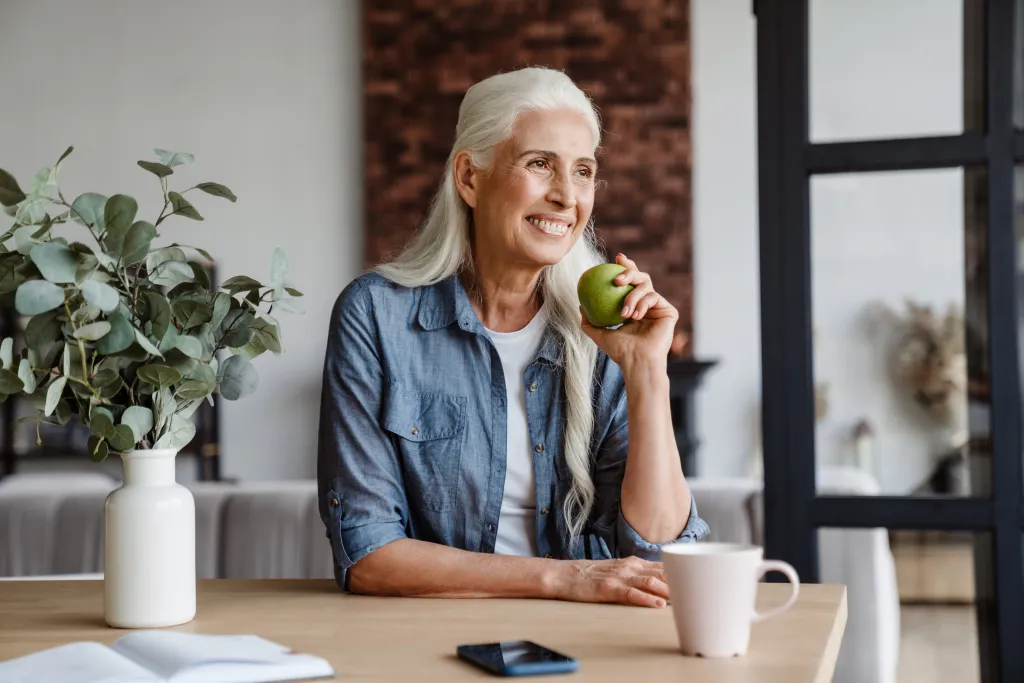 A smiling senior woman sits at a kitchen table, holding a green apple.