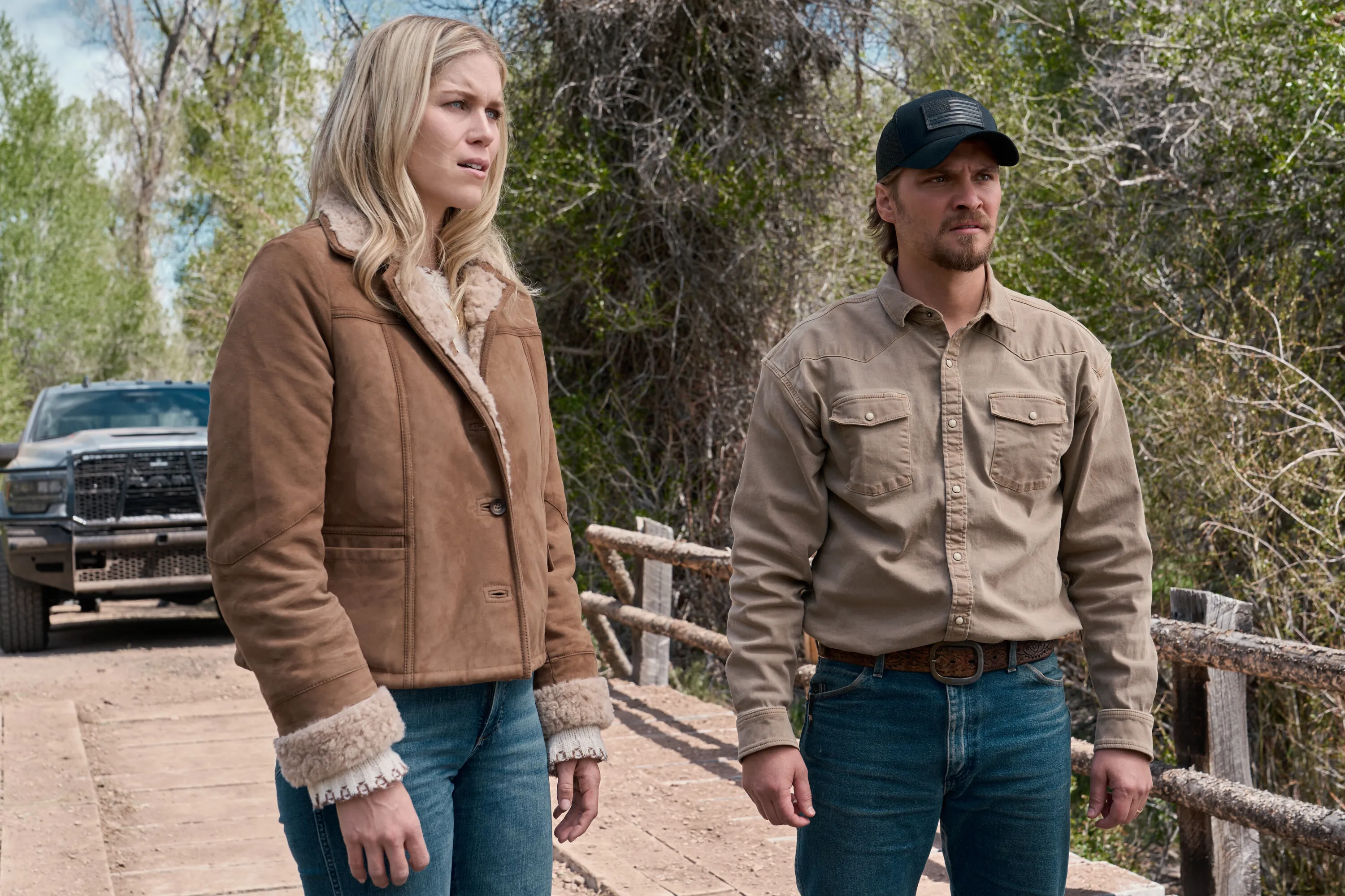 Ellyn Jameson as Dolly Weaver and Luke Grimes as Kayce Dutton outdoors near a truck and wooden bridge.