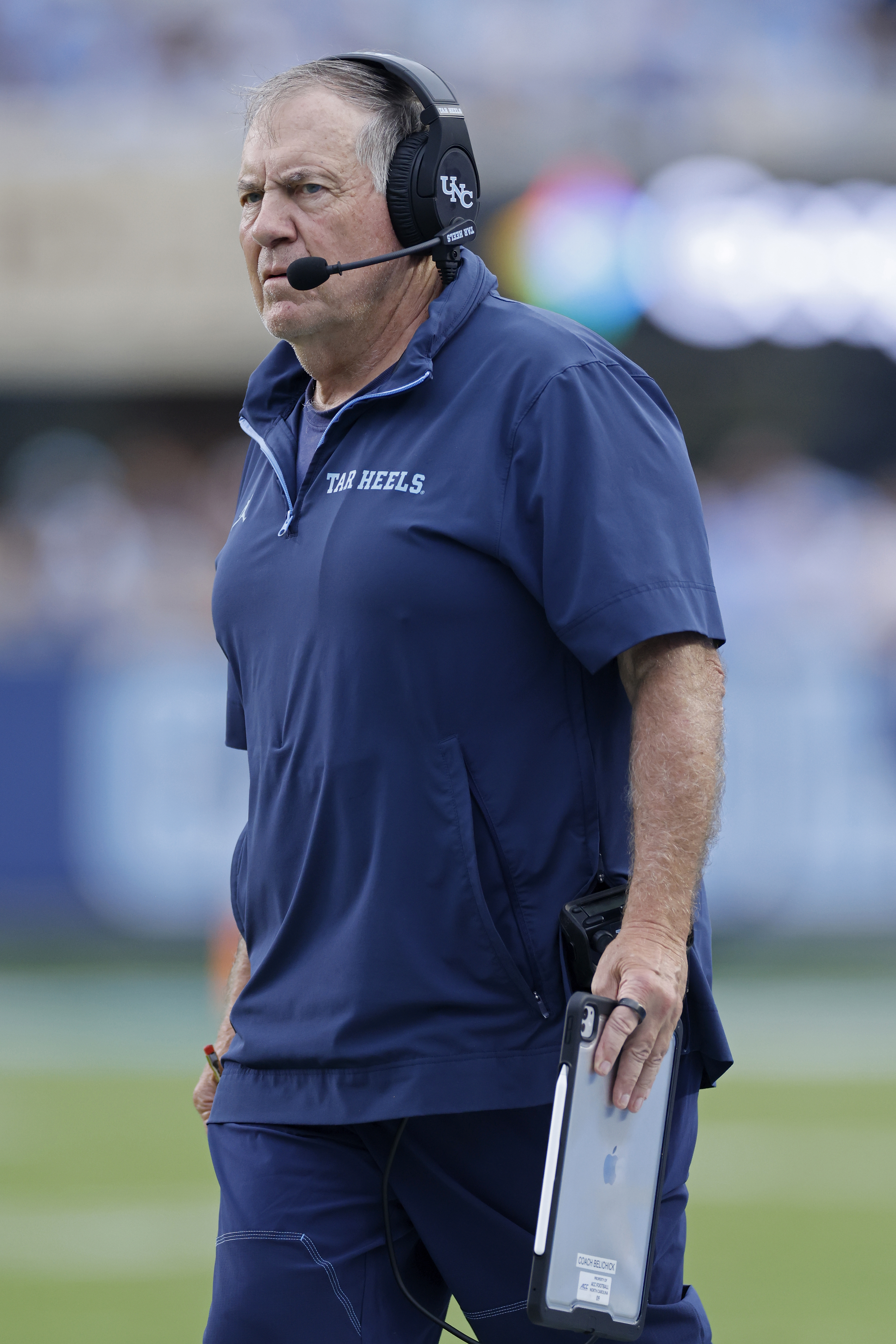 North Carolina Tar Heels head coach Bill Belichick wearing a headset and navy blue pullover while holding a tablet.