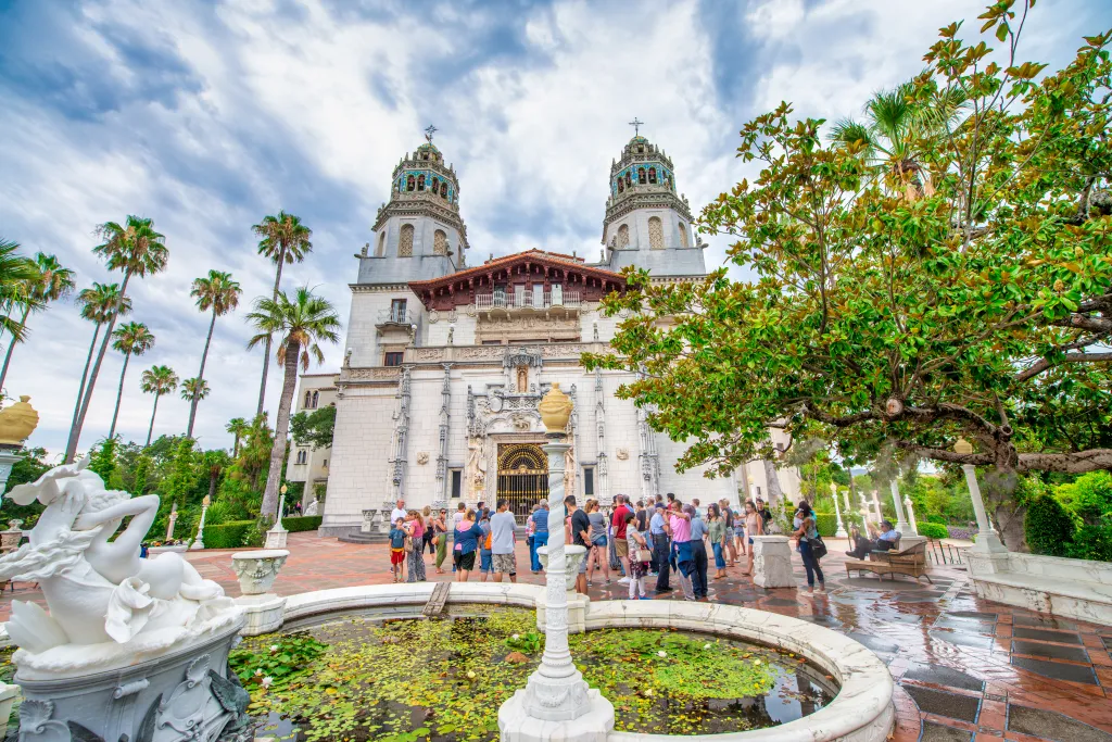 Tourists gather in front of the historic Hearst Castle, featuring ornate twin towers and a plaza with a lily pond.